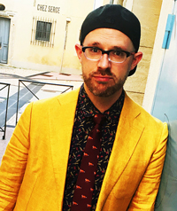 A man in a yellow blazer and glasses looks at the camera.