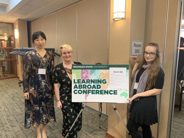 three woman wearing black dresses standing around a sign with green letters saying 'Learning Abroad Conference'