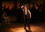 actor standing on a lit up stage, hand on the back of their head