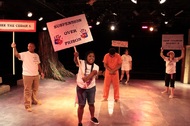 actors protesting with signs, an actor in an orange jumpsuit in looking to the ground in the center