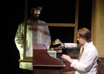 one actor sitting at a desk while another actor stands above him staring