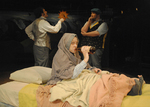 an actor sitting in bed while two actors behind her argue