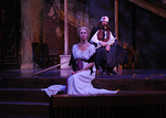 an actor sitting on a stair and looking into the distance, an actor behind her is holding a stick and looking as well