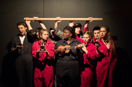 group of actors in jumpsuits posing for a picture, they are holding a stick in the background, the center is holding a ukulele