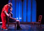 an actor sitting in a chair, another actor is leaning in and grabbing the back support of the chair