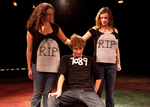 two actors with tombstones plastered on them touch the back of a head of an actor sitting down