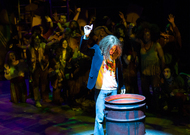 an actor standing with one arm up in front of a oil barrel