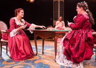 two actors playing cards in big, red dresses, a third actor is watching from a couch