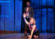 an actor on the ground reaching forward while screaming, an actor stands behind her peering at her