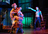 three actors acting as a beast stand on the left side of the screen, an actor on the right, only wearing pants, is pointing a knife in the beast's direction