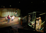 actors standing in the center of stage while two actors on an elevator look down on them