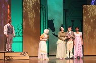 four actors lined up and talking while facing the audience, a male actor is seen standing on the left side of the stage