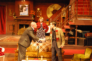two actors standing in front of a couch and shaking hands, another actor is watching behind the couch