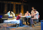 actors talking over a table, two are seated on a chair, one is seated on a bed, and the last one is standing