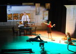 actor standing and holding a wine glass while presenting a projected image of a man to an actor sitting on the ground, there is also an actor passed out on the ground