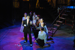 actor with painted face and in a suit is surrounded by actors curiously watching