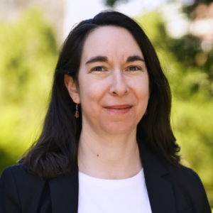 A picture of a woman with long black hair looking at the camera; she has a white shirt and a black jacket.