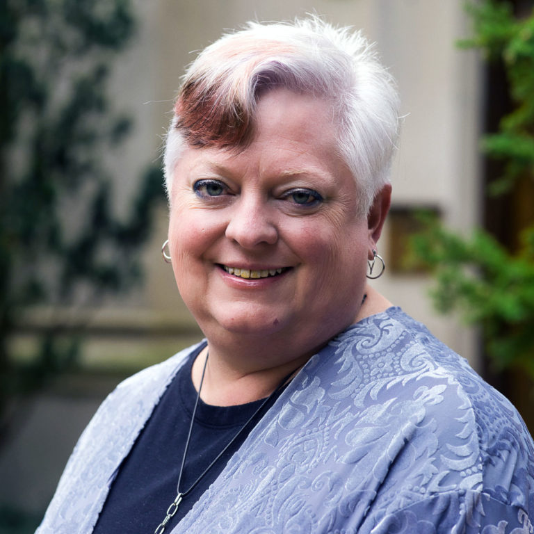 A picture of a woman with short white hair smiling at the camera. She wears a blue patterned sweater and a blue shirt.