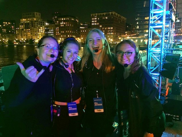 Four women, all wearing headsets, standing outside with a river and skyline in the background. 