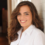 A person with long wavy brown hair, brown eyes, and wearing a white collared button-down shirt smiles against a white brick wall with windows in the background