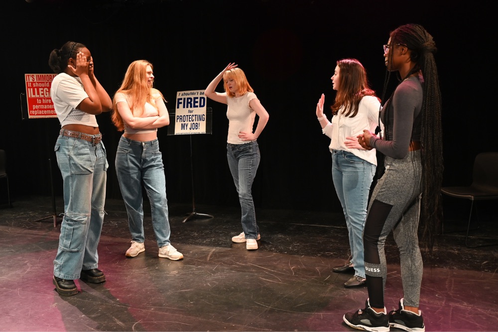 five female actors on stage with a sign in the background that says: "Fired for Protecting My Job!"