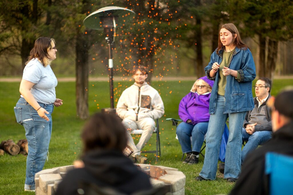 Two individuals stand and perform near a fire pit outdoors, with orange embers rising in the air. They are surrounded by seated spectators in lawn chairs, dressed for cool weather, under trees and portable heaters.