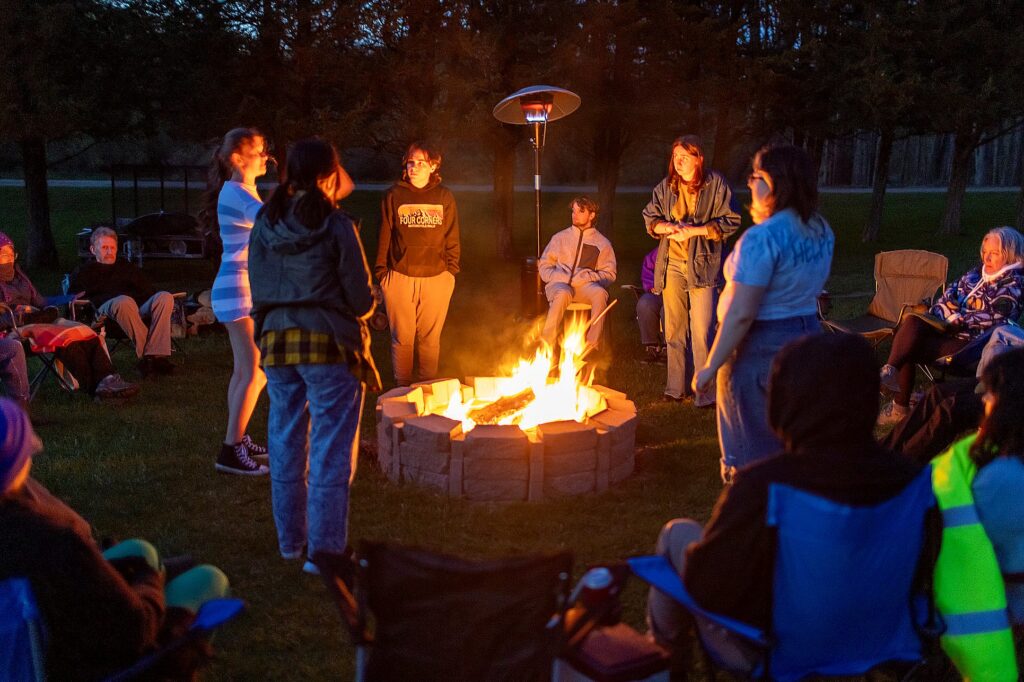 A group of people stands around a fire pit at night, warmly lit by the flames. Others sit in a circle of camp chairs watching attentively. The gathering takes place outdoors on a grassy field, surrounded by trees and illuminated by a tall outdoor heater.