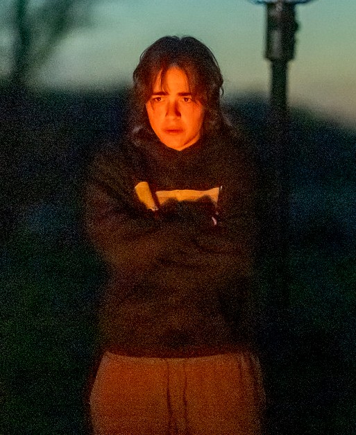 A person wears a dark hoodie and loose pants, arms crossed over their chest, outdoors at dusk or nighttime, lit from below by a warm light source