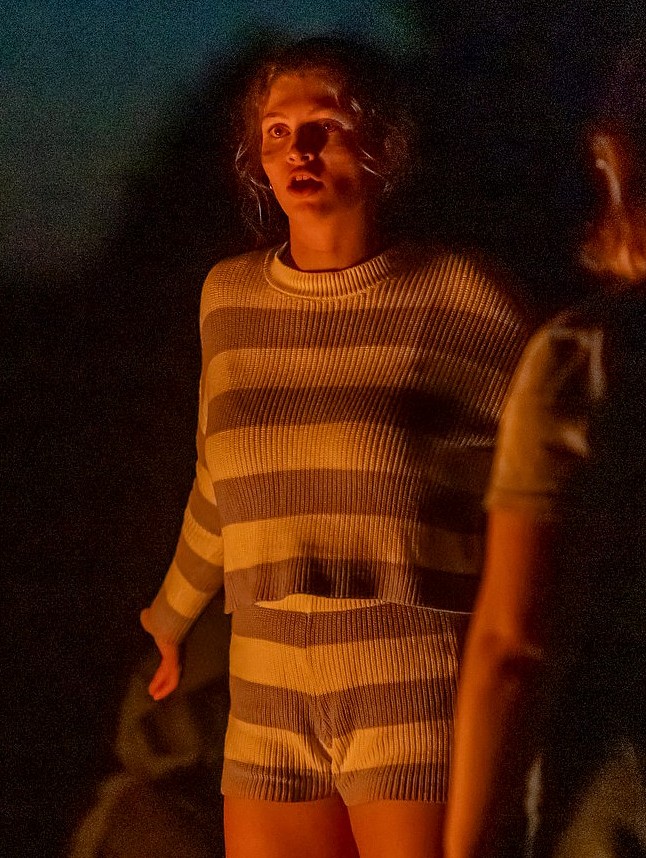 A person in a striped, long-sleeved sweater and matching shorts is lit dramatically from below by a fire.