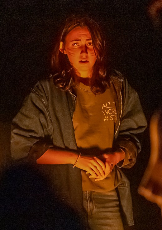 A person stands in a spotlight of warm light, wearing a dark denim jacket over a shirt that reads "ALL THE WORLD'S A STAGE.