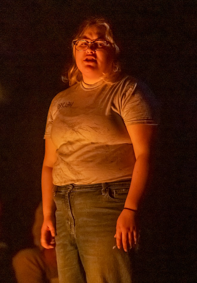 A person wearing glasses and a light-colored T-shirt stands in a warm glow against a black background.