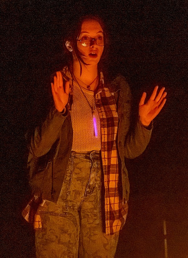 A person stands in front of a dark background, illuminated by a warm, orange light. They wear glasses, a layered outfit with a plaid shirt and denim jacket, and a pendant around their neck.