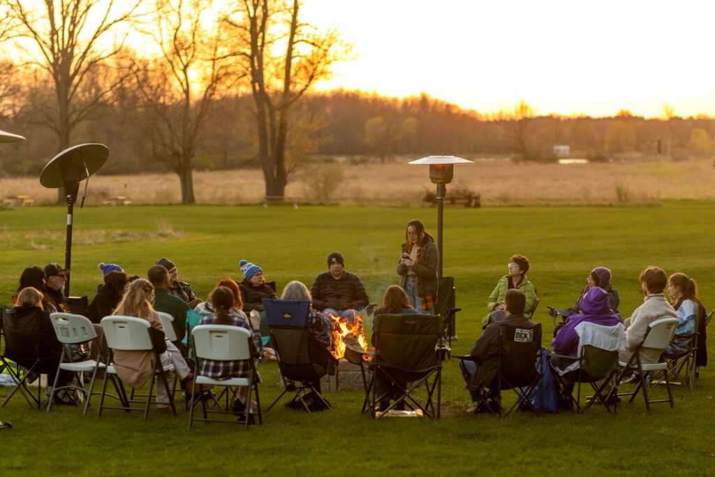 A large circle of people sits in folding chairs around a fire pit on a grassy field at sunset. One person stands and speaks while others listen, with trees and open landscape stretching into the distance.