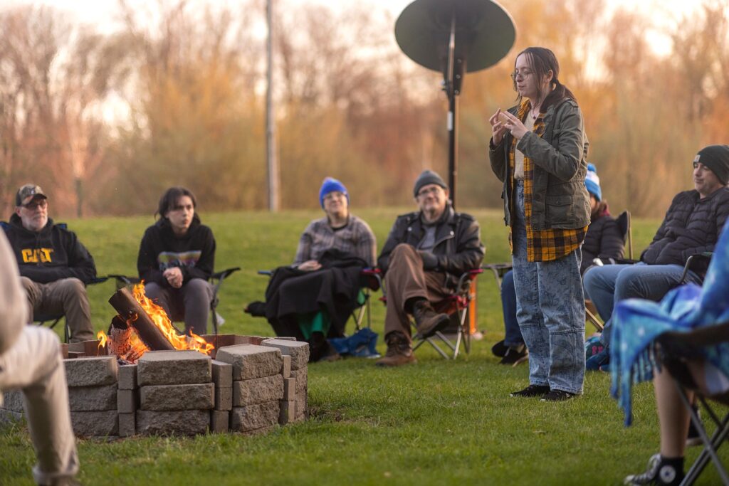 People wearing coats and hats sitting in folding chairs around a fire pit on a grassy field. One person stands and speaks while others listen, with trees in the distance.