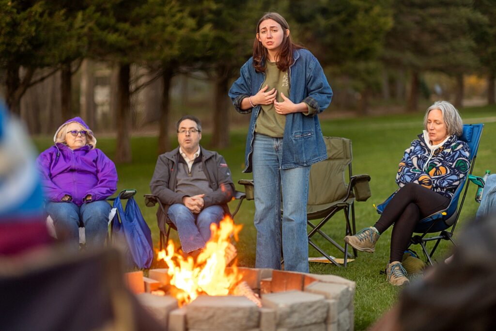 A personstands and speaks near a fire pit, using hand gestures, while others seated nearby listen attentively. The group is gathered in lawn chairs on a grassy field with trees in the background, dressed warmly for the evening.