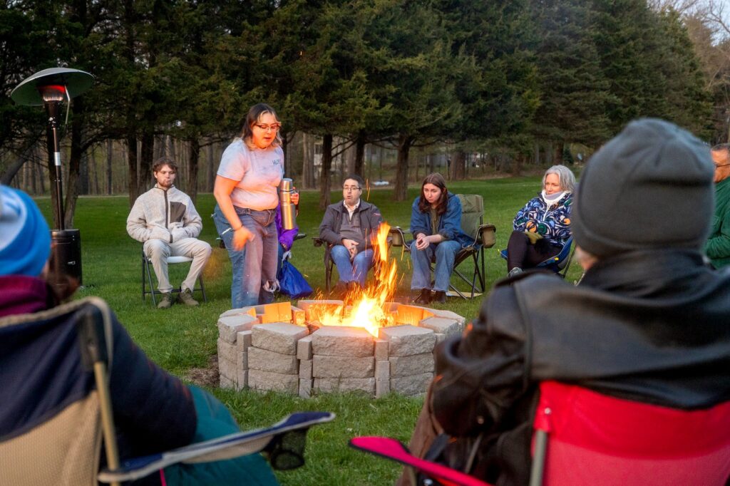 A person stands near a glowing fire pit holding a thermos, speaking to a circle of people seated in camping chairs. The group is gathered on a lawn with a forested background, dressed for cool weather as dusk falls.