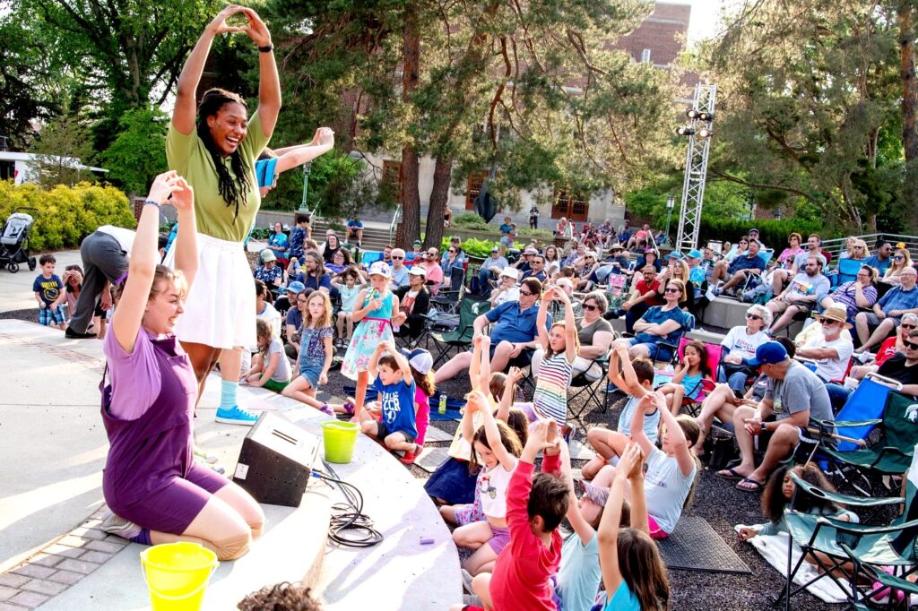 A lively outdoor theater scene where two performers lead a group of children in interactive movements. The crowd includes families and children, many of whom are mimicking the performers' arm movements. The setting is sunny, with trees, a campus building, and stage lighting in the background.