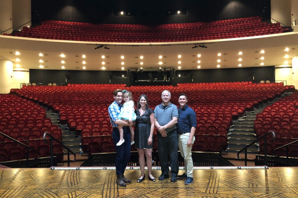 A photo of five individuals standing on an empty theater stage, smiling at the camera with the expansive, red-seat auditorium stretching behind them