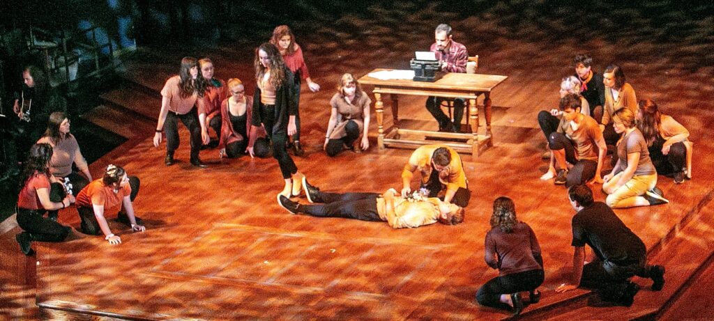 A theatrical performance on a stage with dramatic lighting. Several actors are grouped around two performers, one lying down and another attending to them. A desk and typewriter are positioned prominently in the background with another performer sitting at the desk.