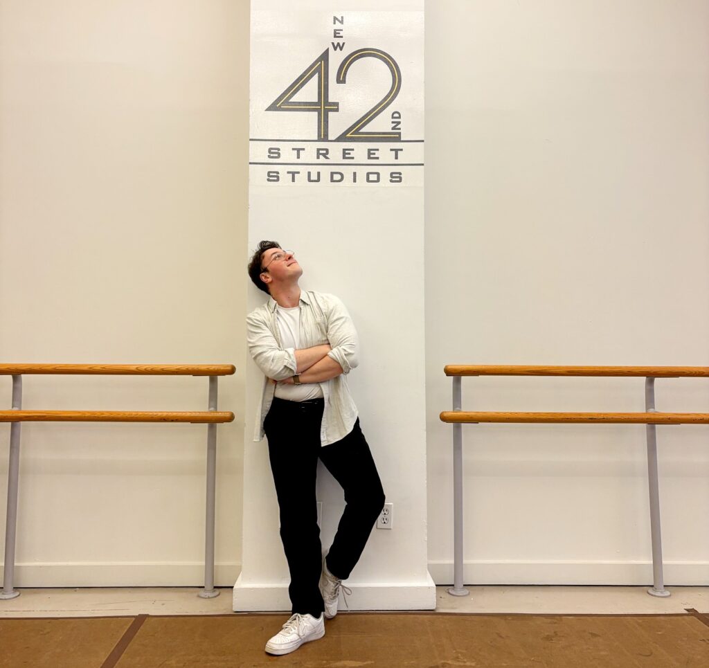A person leaning against a pillar marked 'New 42 Street Studios.' They wear a casual white shirt and black pants, looking upward.