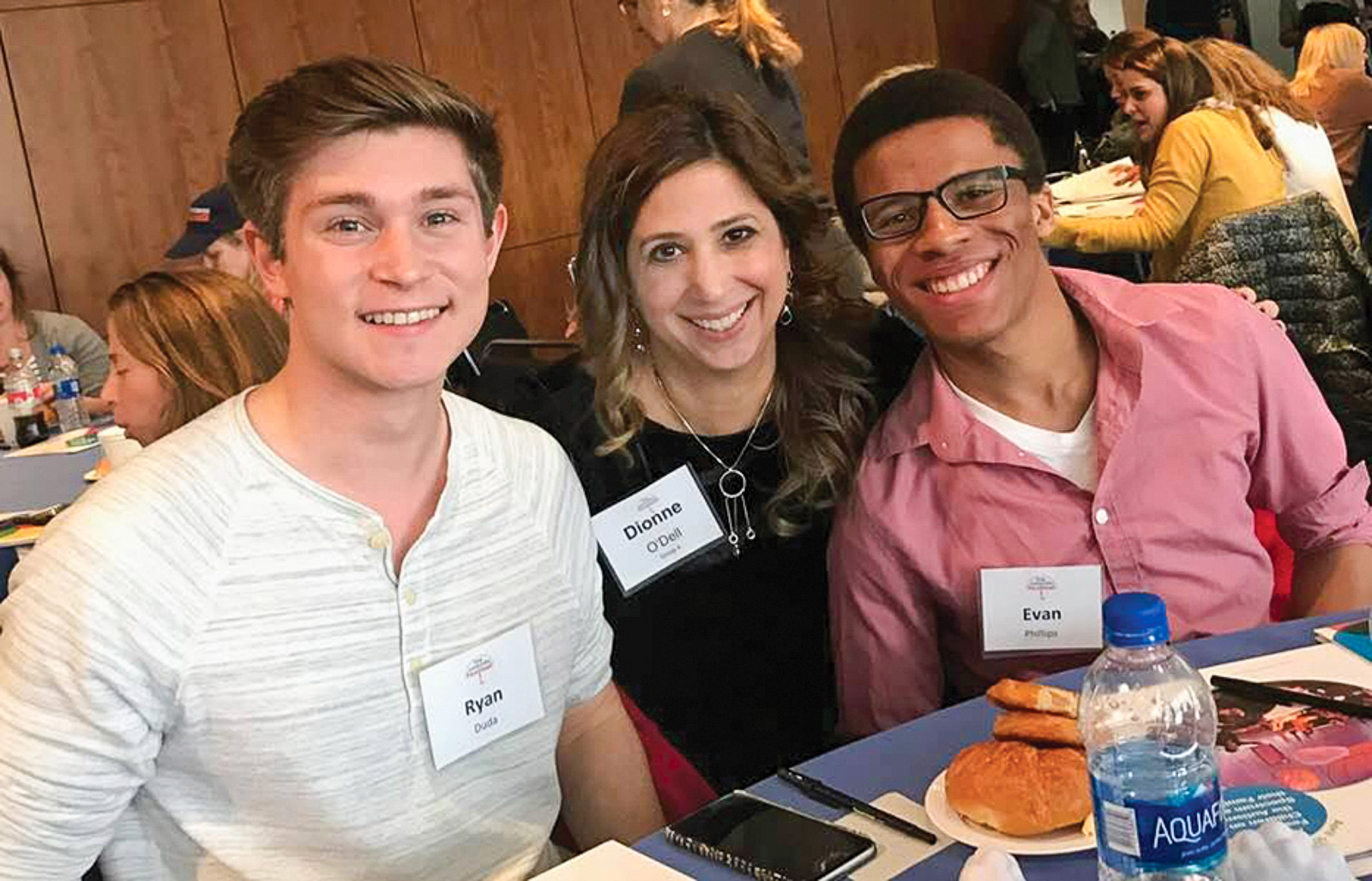 Three people sit closely together at a round table, smiling at the camera in a crowded indoor event space. The table is set with food, water bottles, and event programs.