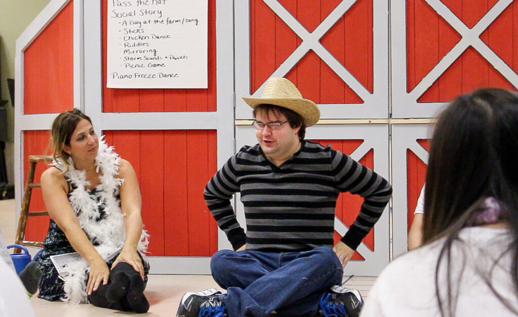 A woman in a feather boa sits on the floor next to a person in a cowboy hat, both in front of a red and white barn-themed set. Others are partially visible in the foreground, seated and watching.