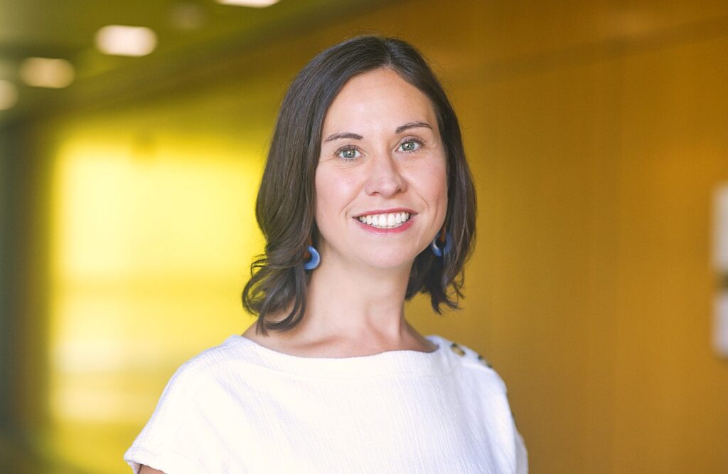 A woman with shoulder-length dark brown hair smiles while standing in front of a softly blurred, golden-yellow background. She wears a textured white blouse with button details on the shoulder and blue statement earrings.