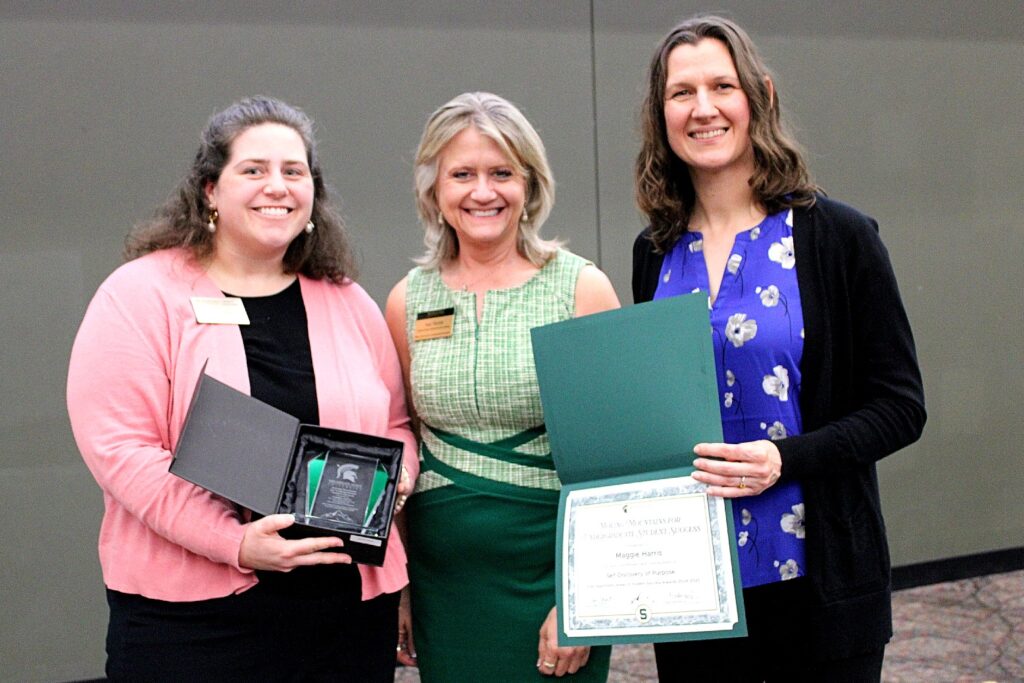Three women pose at an award ceremony. The woman on the left wears a black top with a pink cardigan and holds an open award box. The woman in the center wears a green plaid dress with a dark green waistband. The woman on the right wears a royal blue blouse with white flowers and a black cardigan. She holds a certificate awarded to Maggie Harris for “Self-Discovery of Purpose.”