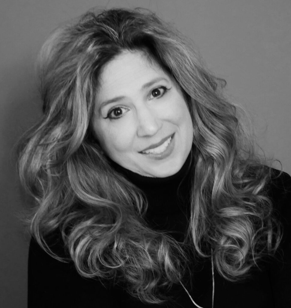 A black and white portrait of a woman with long wavy hair, smiling slightly while wearing a dark turtleneck against a plain background.