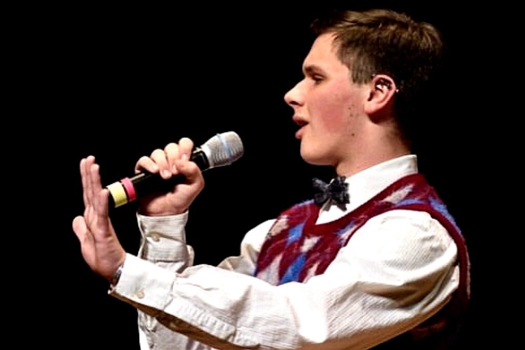 A photo of an individual performing on stage, holding a microphone and wearing a patterned sweater vest with a bowtie. The background is dark, emphasizing the performer's focused expression.