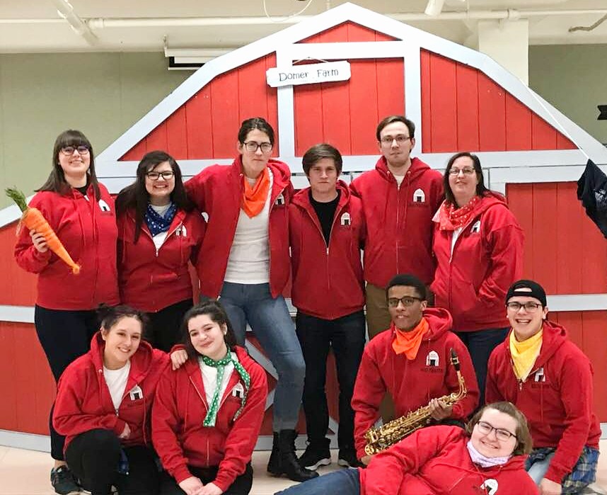 Eleven people all wearing red hooded sweatshirts standing and sitting in front of a red barn theatre set and on the barn there is a sign that says "Domer Farm."