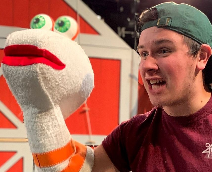 Man wearing a green baseball hat backwards and a maroon T-short. He is performing with a puppet on his right hand.