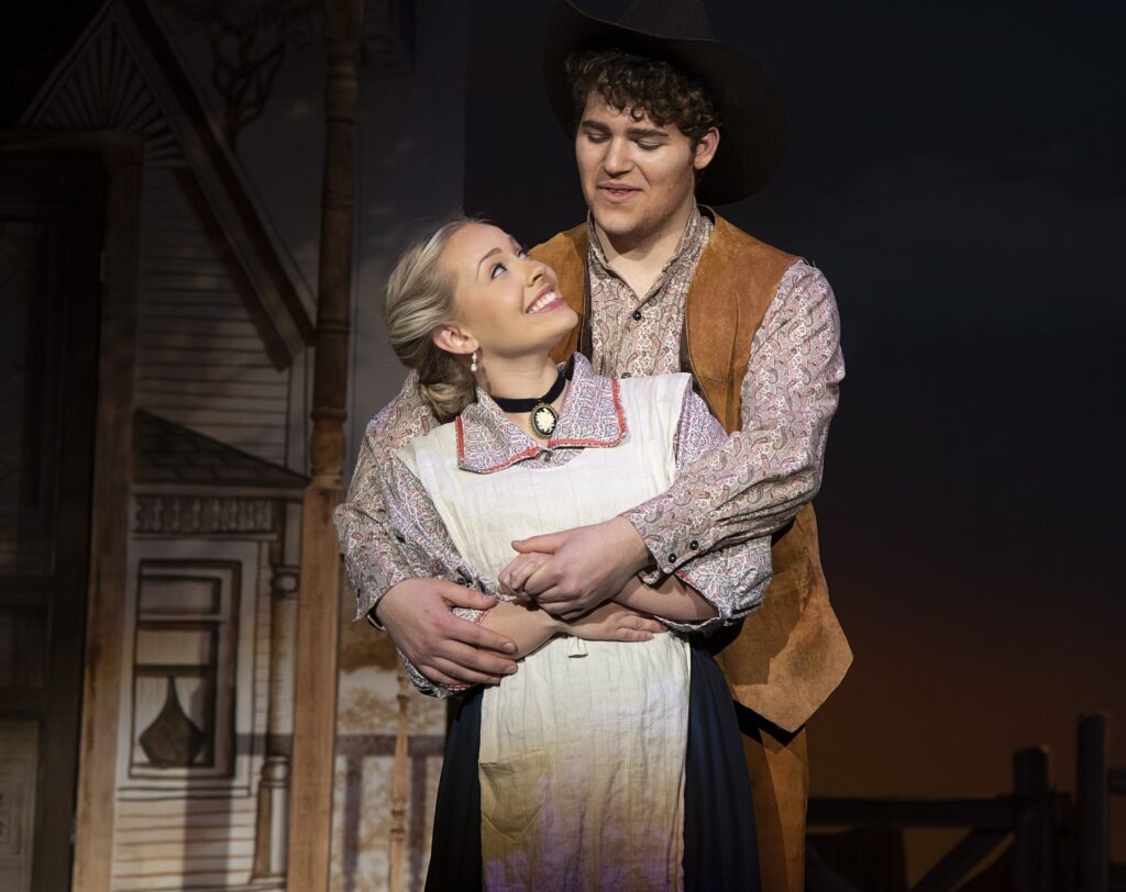 A young woman and man perform on stage. The woman, wearing a long dress with a white apron and a cameo brooch, smiles warmly while looking up at the man behind her. The man, dressed in a Western-style outfit with a suede vest and cowboy hat, holds her gently.