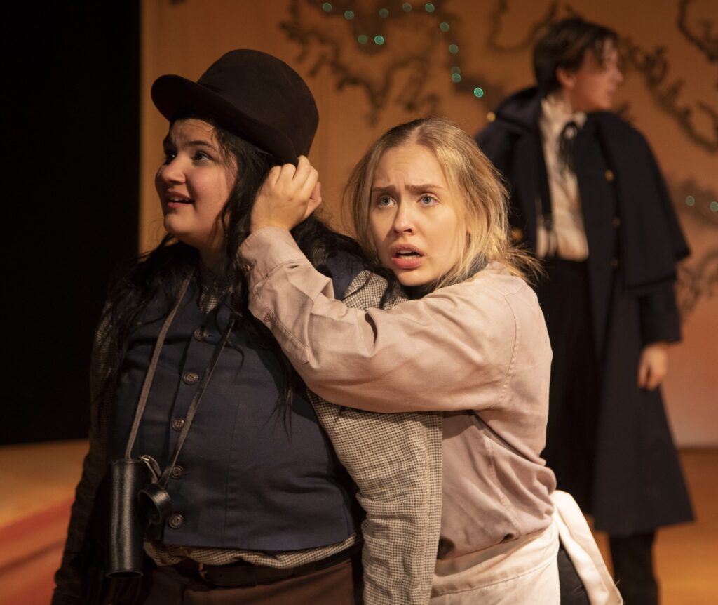 Two actors perform a tense moment on stage. One, wearing a bowler hat and vest, looks offstage. The other, dressed in a light shirt and apron, clutches the first actor’s arm with a worried expression.